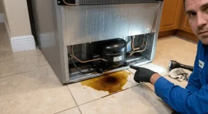 A close-up of an LG refrigerator compressor with a hand pointing to a dark, viscous oil pool on a tile floor, with a technician ID card visible. This image demonstrates one of the final signs of a failing linear compressor: pooling oil under or behind the appliance.